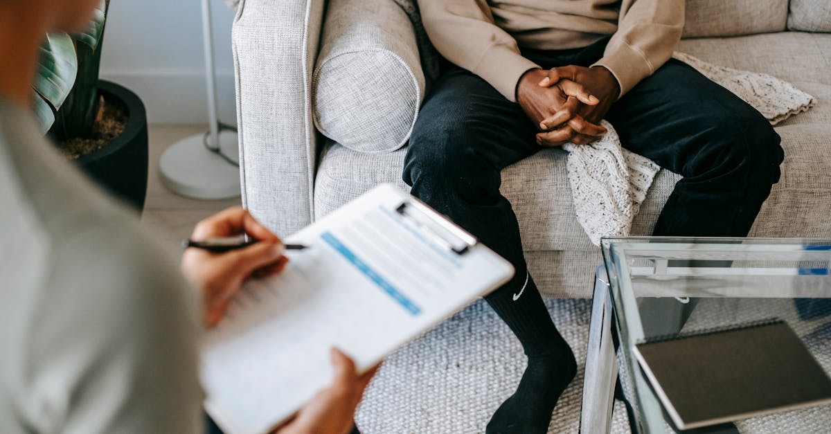 Blurred unrecognizable female therapist taking notes on clipboard while sitting in front of ethnic male patient with clasped hands during meeting