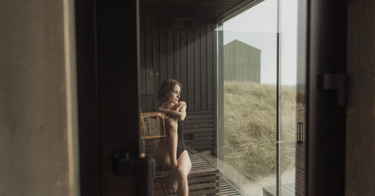 A woman in a bikini relaxes inside a cozy wooden sauna with a view of grassy dunes outside.
